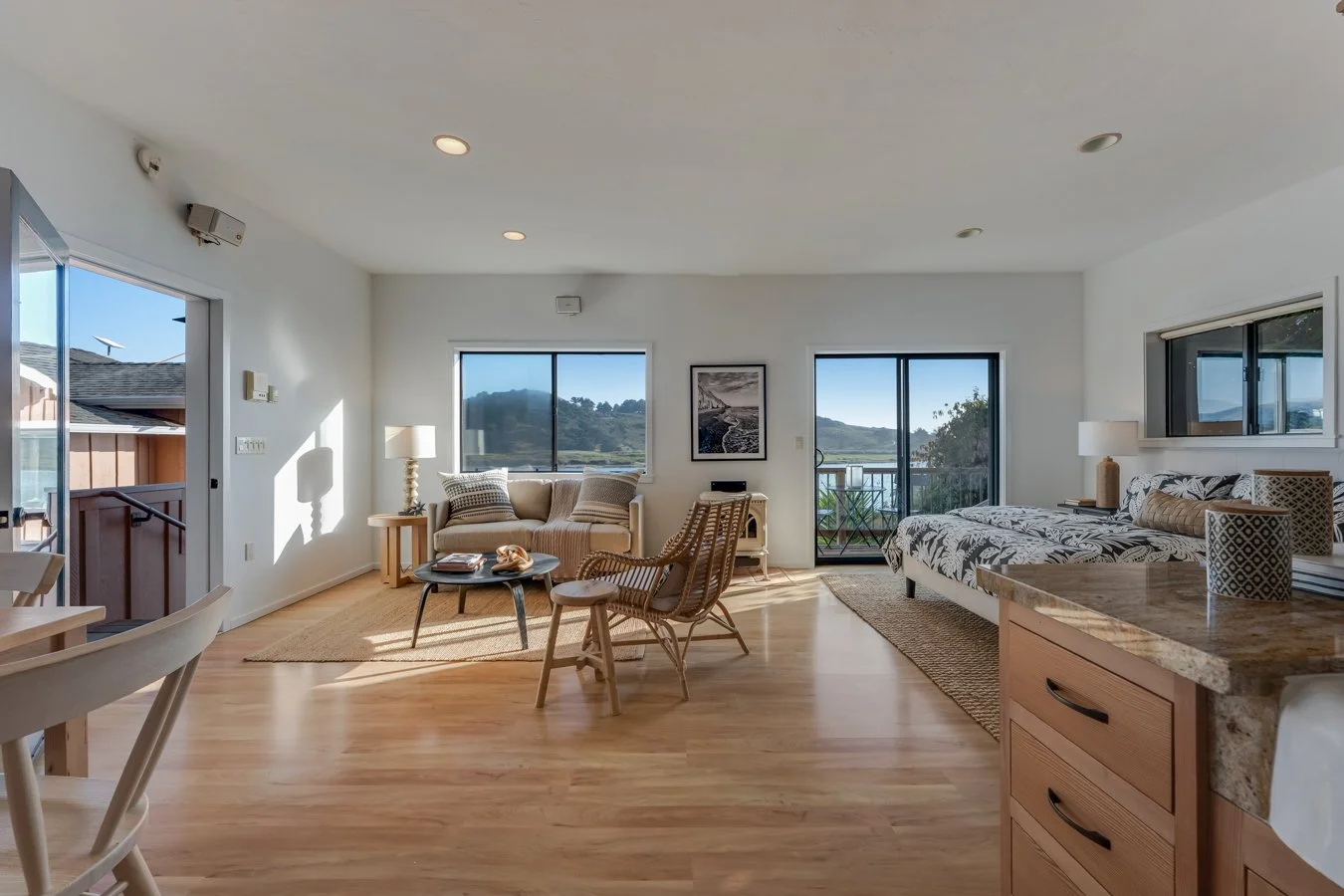 Living room with a beige sofa, rattan chair, and a bed with patterned linens, large windows and sliding doors showing outdoor views, hardwood floors, ceiling lights, and a kitchen counter with mugs.