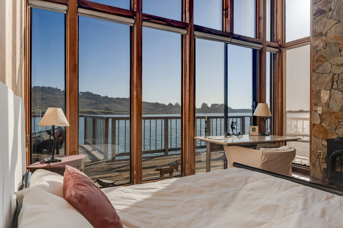 Room with large floor-to-ceiling windows overlooking a lake, with a bed in the foreground and a small desk with a lamp and decorative items near the windows.
