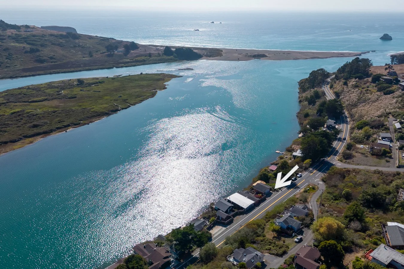 Aerial view of a coastal area with a waterway running parallel to the ocean, houses along the road, and a white arrow pointing to a house.