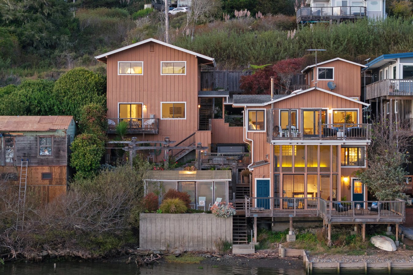 Multi-story waterfront homes on a hillside at dusk, with outdoor decks and windows illuminated from inside.