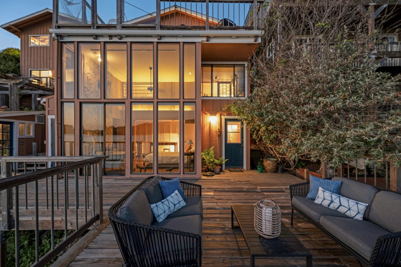 A wooden deck outdoor patio with modern furniture, including two gray sofas with blue pillows and a black coffee table with a lantern, in front of a multi-story home with large glass windows and a warm interior lit by yellow lighting.