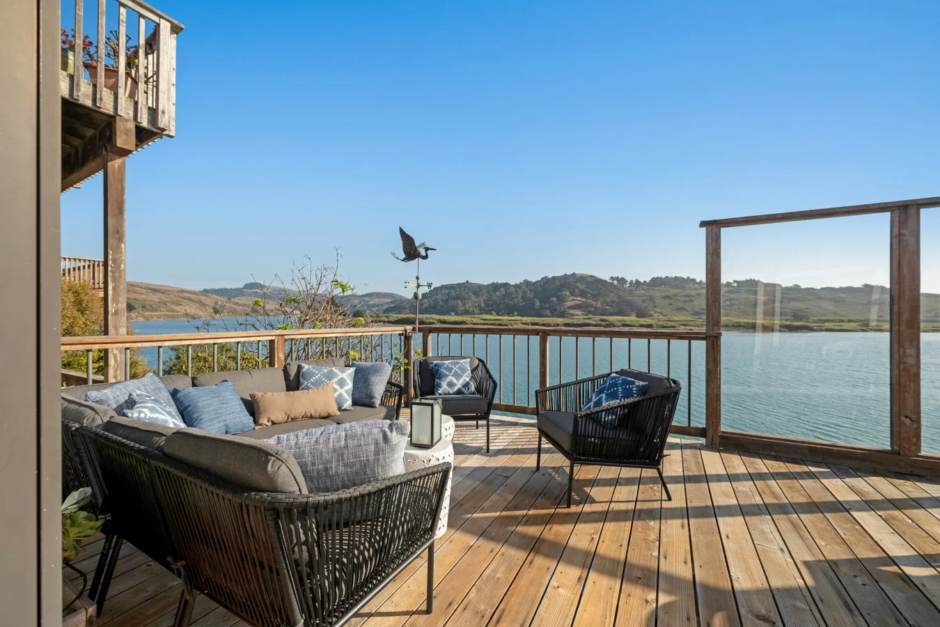 Outdoor wooden deck with seating area, overlooking a lake and distant hills, under a clear blue sky, with a weather vane and bird in flight.