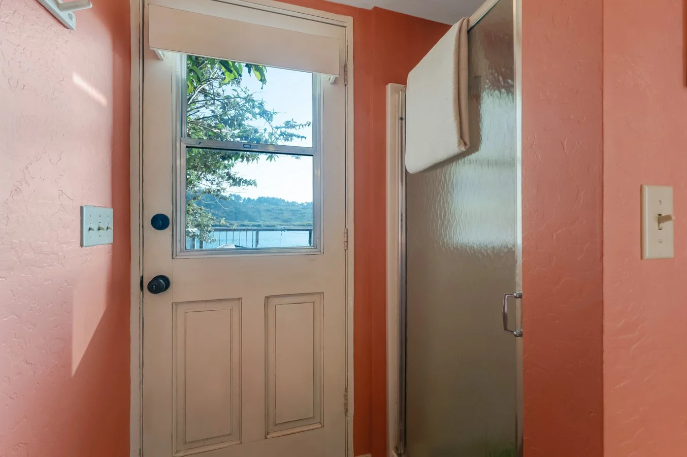 Interior view of a small room with peach-colored textured walls, a white door with a window showing an outdoor landscape, and a metal shower stall with a towel hanging on top.