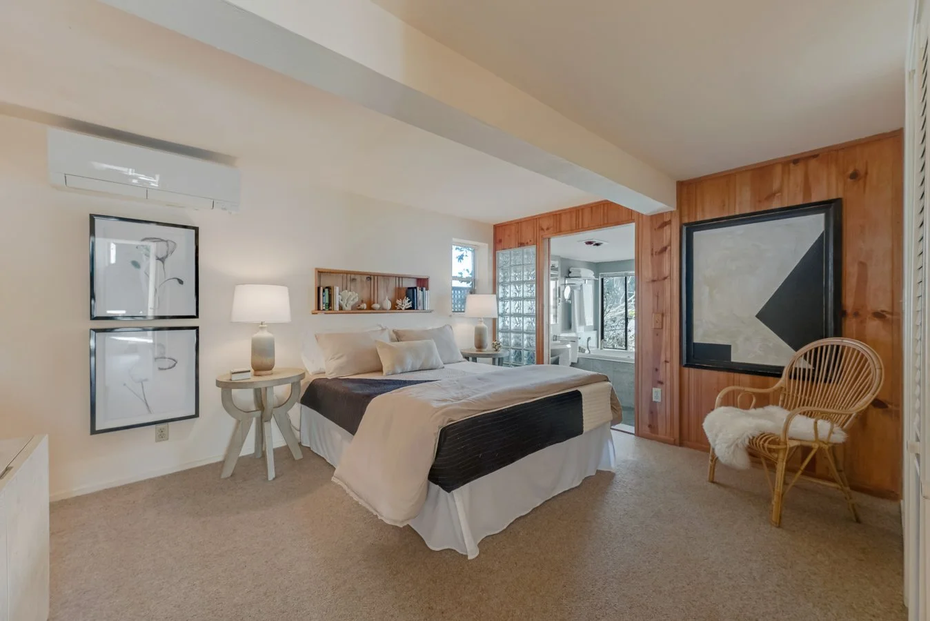 A cozy bedroom with a large bed with white and beige bedding, two night lamps on either side, wooden wall paneling, wicker chair with a white blanket, and framed artwork on the walls. There is a bathroom visible through an open door.