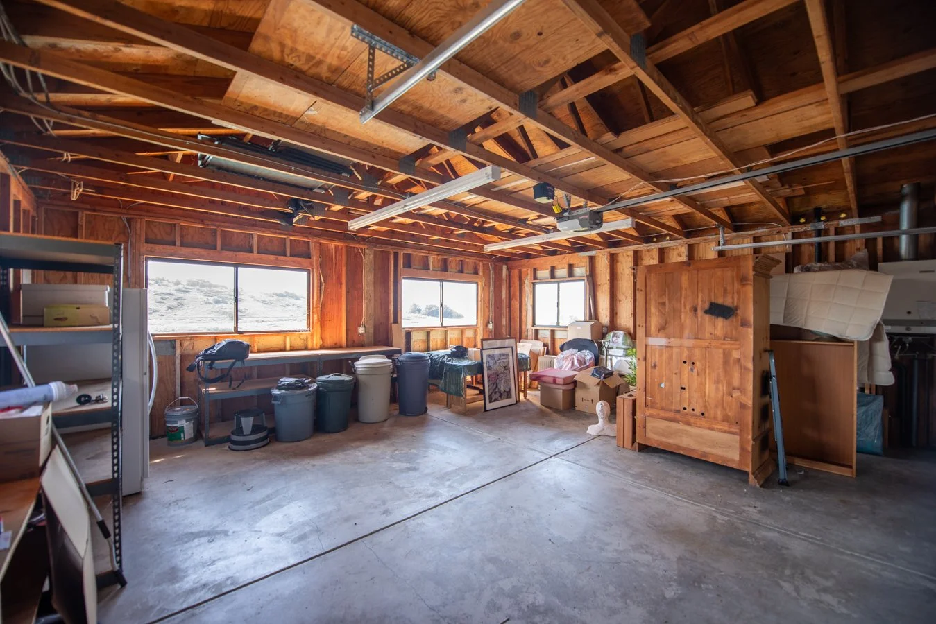 A cluttered garage with wooden walls and ceiling, multiple windows, various storage bins, framed pictures, a lamp, and miscellaneous items.
