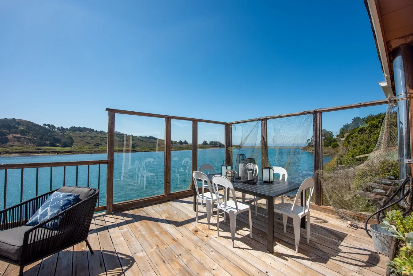 Outdoor deck overlooking a lake with a wooden railing, white chairs, a black table with lanterns, and a cushioned chair with a pillow. Beyond the deck, there is a body of water and rolling hills under a clear blue sky.