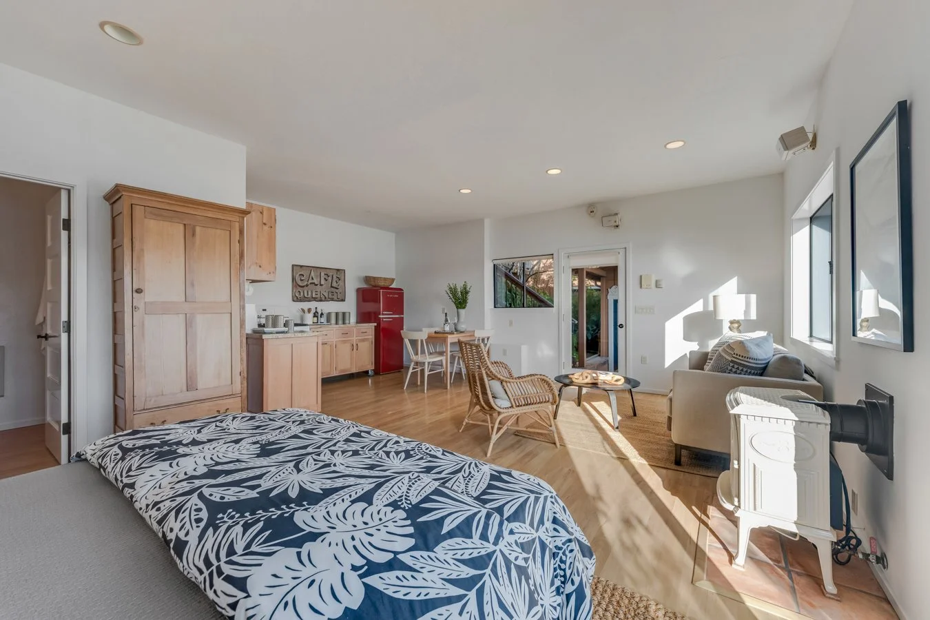 Open-plan living room and kitchen with a bed in the foreground, a wooden wardrobe, a red refrigerator, a dining table with chairs, a sitting area with a sofa and armchairs, and a door leading outside, all illuminated by natural light.