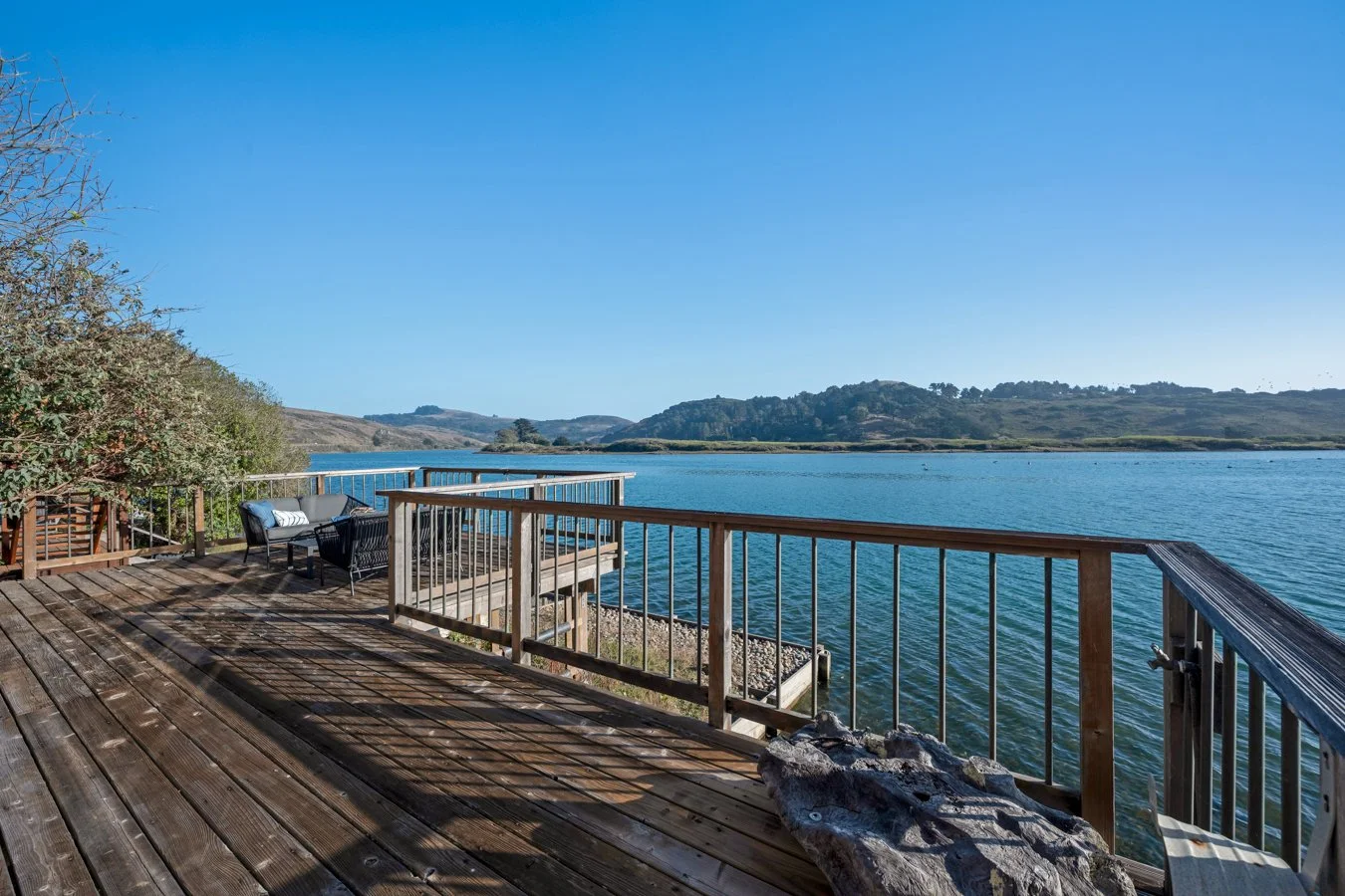 A wooden deck overlooking a large body of water with hills in the distance, a black outdoor sofa with a white cushion, and trees on the left side.