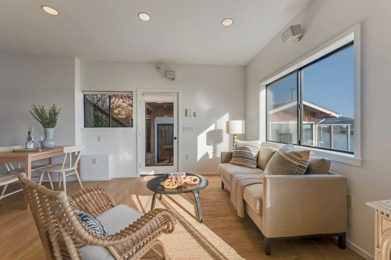 Living room with a beige sofa, large window, rattan armchair, round coffee table with decorative objects, dining table with chairs, hardwood floors, and wall-mounted lights.