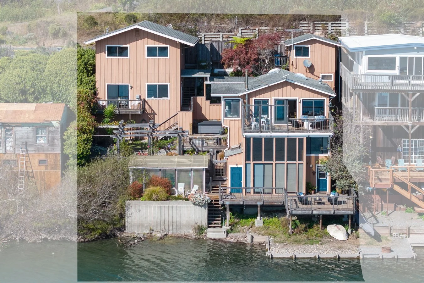 A multi-story house built on a lakeshore with decks, stairs, and a boat dock, surrounded by trees and other houses.