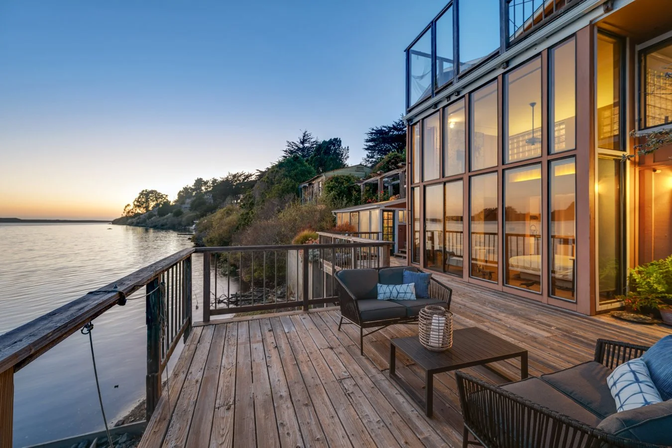 A wooden deck overlooking a calm body of water during sunset, with outdoor furniture and a modern glass house reflecting the warm interior lighting.