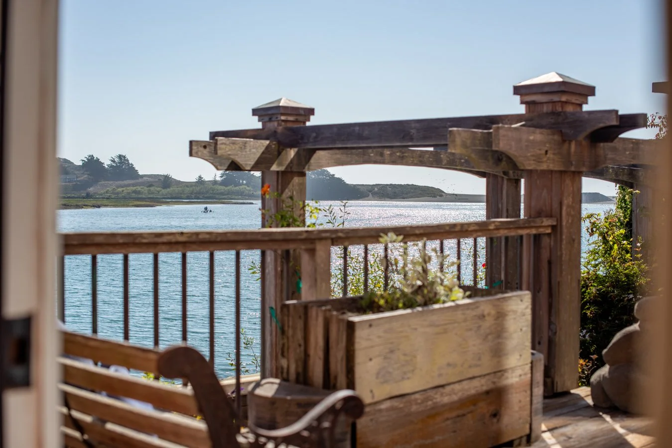 View of a calm body of water with a small boat in the distance, seen from a wooden porch with a railing and a bench, during daytime with clear sky.