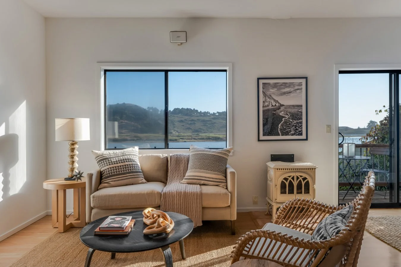 Living room with beige sofa, rattan armchair, black coffee table with books and art sculpture, side table with lamp, wall art, and sliding glass door leading to balcony with outdoor view.