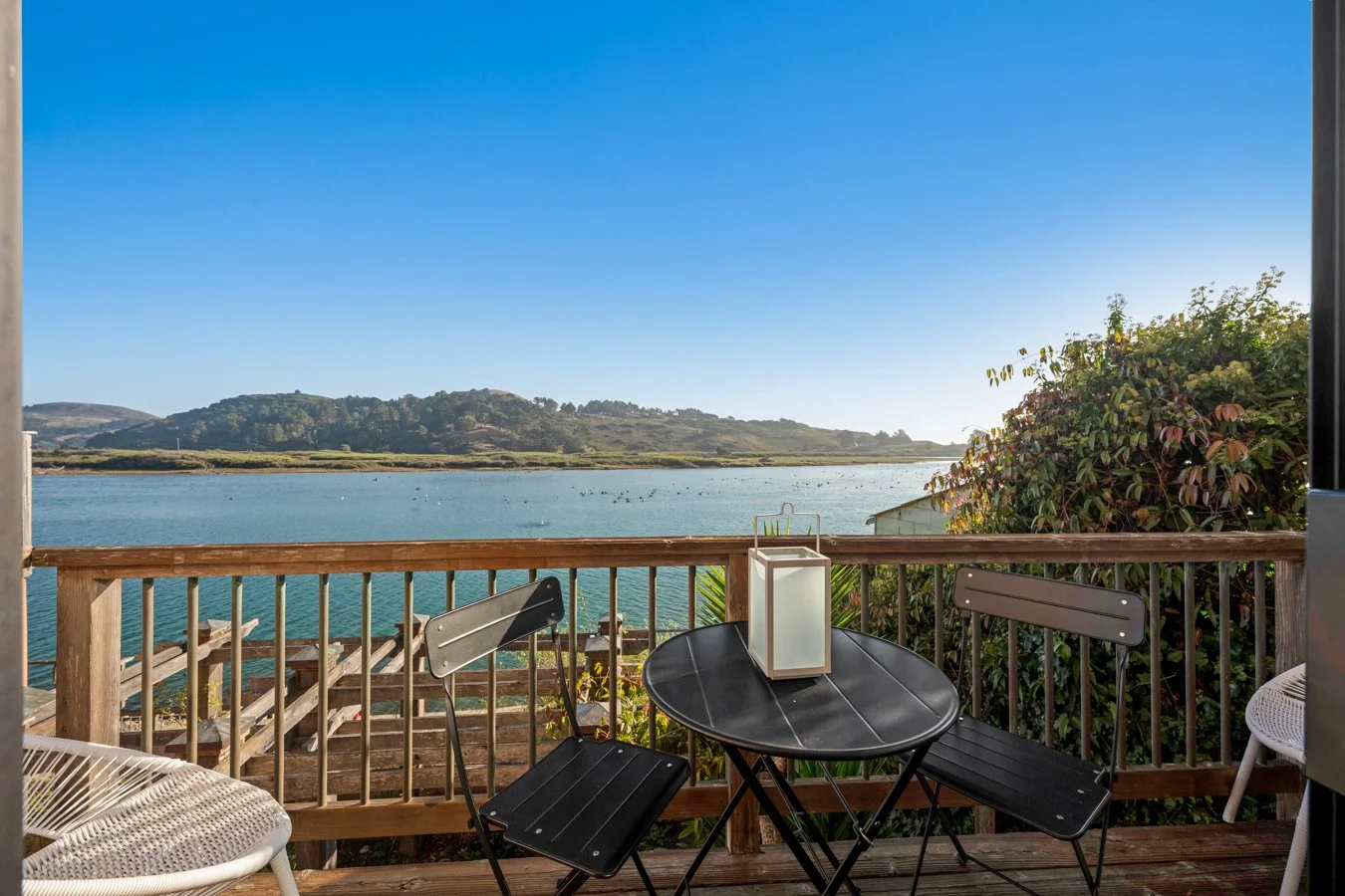 A wooden deck overlooking a body of water with hills in the distance; outdoor furniture includes a small black table with a white lantern, two black chairs, a white wicker chair, and a white chair partially visible on the right; lush greenery on the 