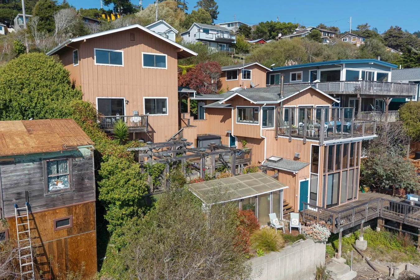 A hillside view of multiple homes with wood and glass exteriors, surrounded by trees and greenery, under a clear blue sky.