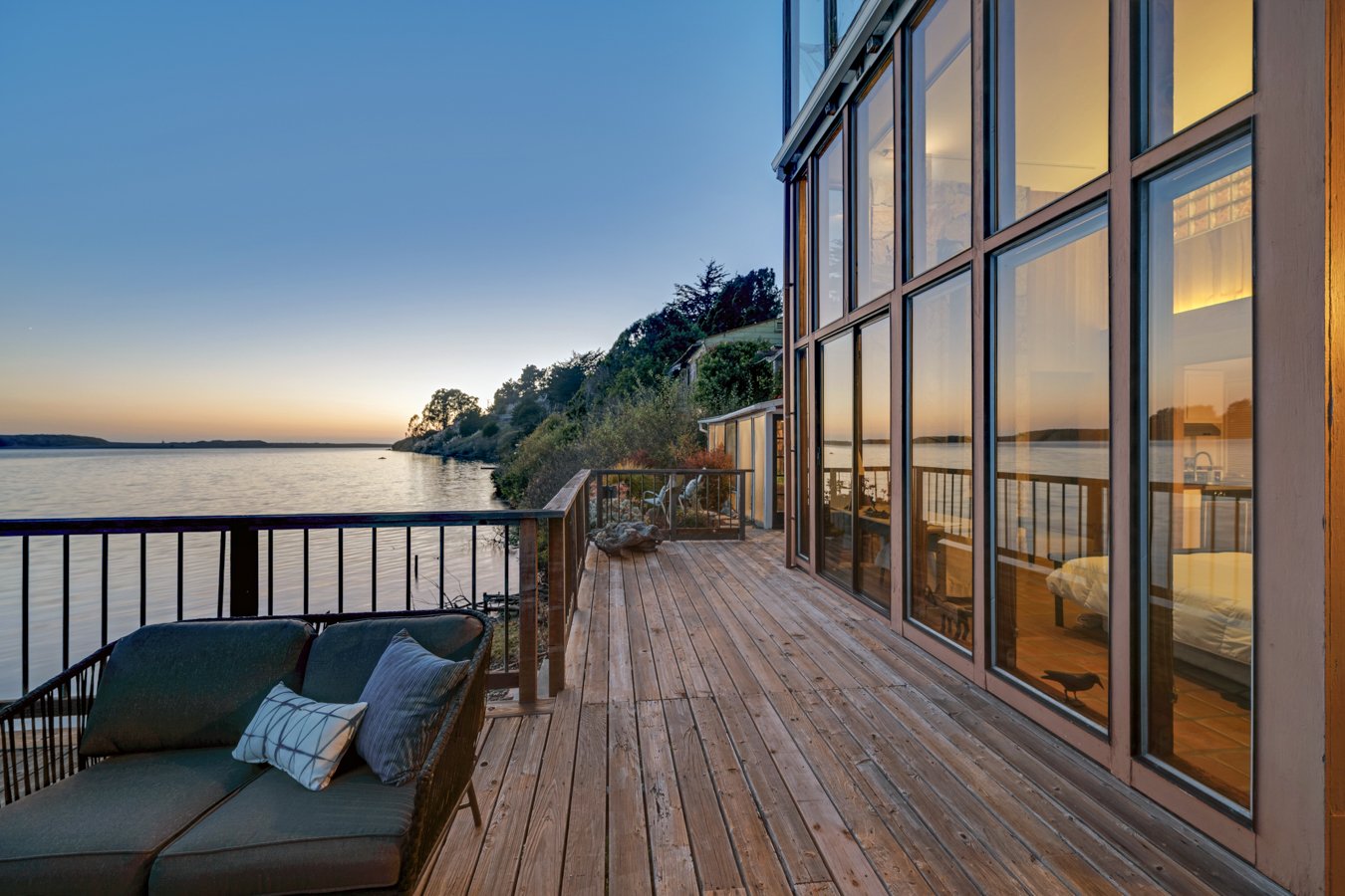 A wooden deck overlooking a body of water, with glass windows on a building reflecting the sunset, and outdoor furniture including a cushioned lounge with pillows.
