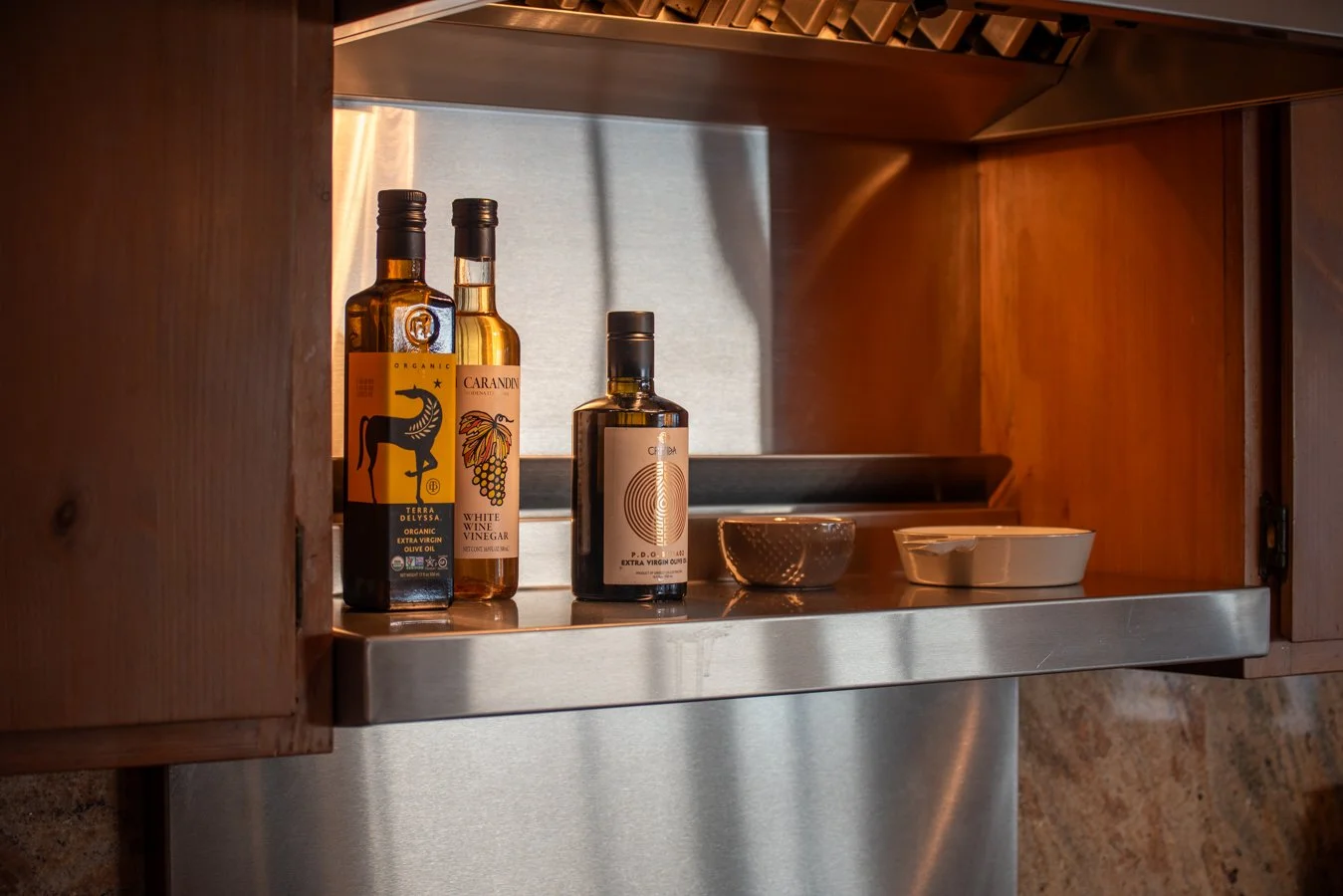 Three bottles of olive oil and vinegar bottles, a small bowl, and a baking dish on a kitchen shelf with wooden cabinets and a stainless steel surface.