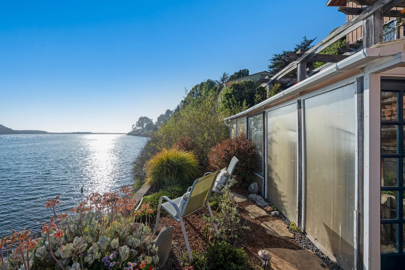 A lakeside outdoor patio with chairs, plants, and a greenhouse, overlooking a body of water during sunset or sunrise.