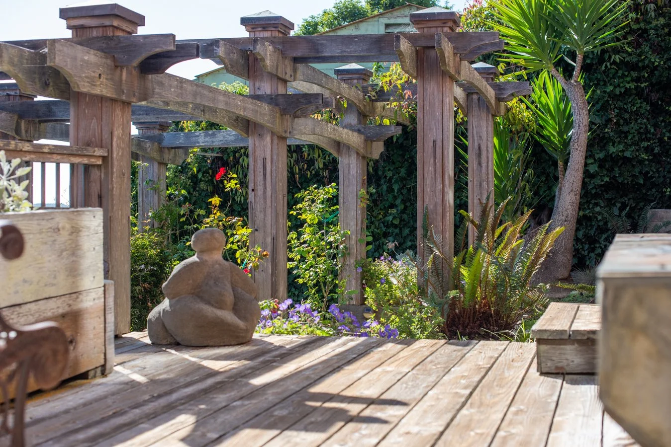 Garden with wooden deck and pergola, featuring a stone figurine, lush green plants, and colorful flowers.