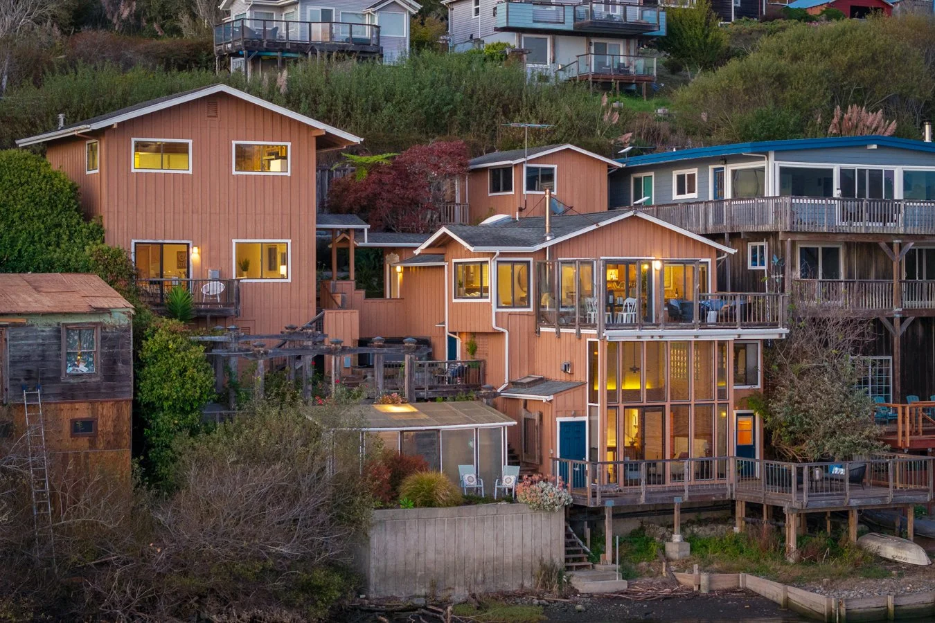 Multiple houses built on a hillside with a variety of wooden decks and balconies, some with outdoor furniture, and lush green bushes and trees surrounding the area.