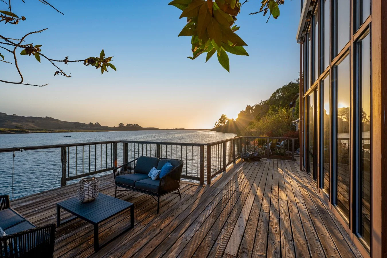 Sunset view from a wooden deck overlooking a river with trees on the horizon, patio furniture including a black sofa with blue cushions, a coffee table, and a small lantern.