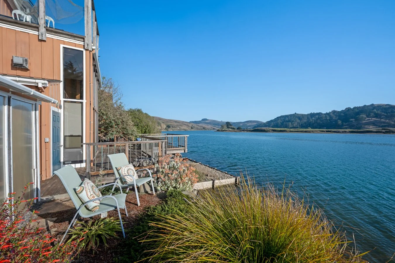 Beach house with outdoor chairs near water and hills in the background