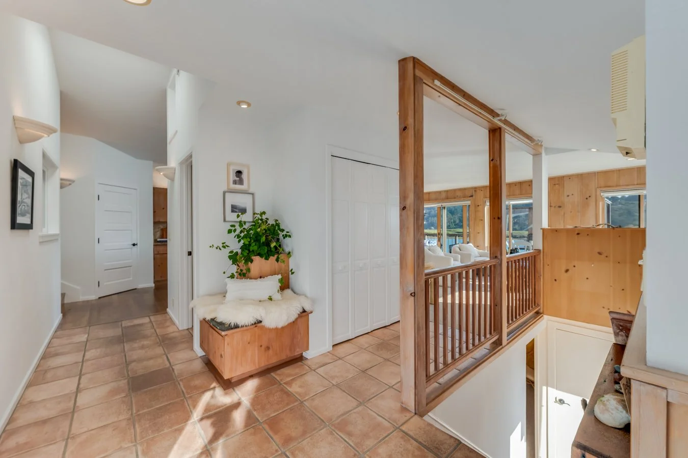 Interior view of a cozy home with terracotta tiled floor, white walls, wooden accents, and large windows showing outdoor scenery. There is a small wooden bench with white cushions and a plant, a hallway leading to a kitchen, and a partially visible s
