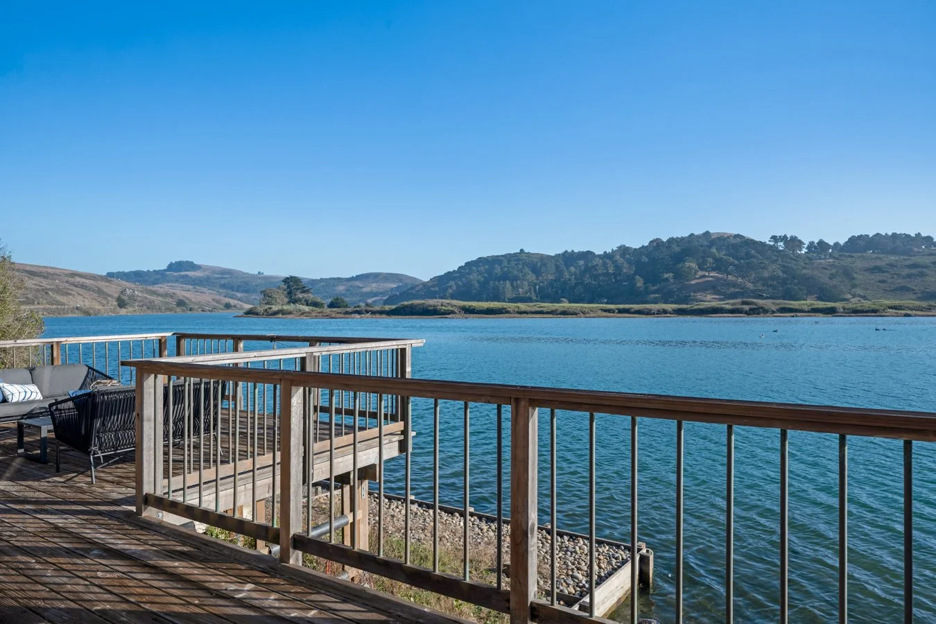 A wooden deck with outdoor furniture overlooking a calm blue lake, with hilly green land in the background under a clear blue sky.