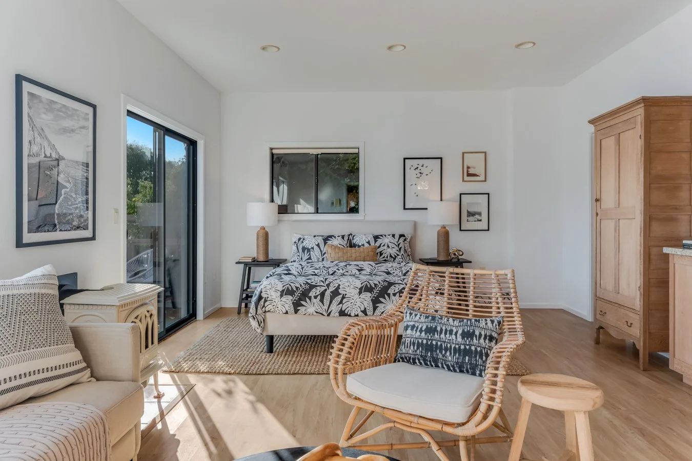 Bright living room with a bed, rattan chair with cushion, framed black and white artwork, sliding glass door, and wooden armoire.