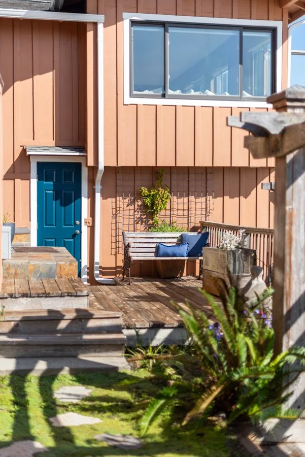 Exterior view of a house with a wooden deck, a blue door, and a large window. There is a bench with cushions, a potted plant, and some garden plants in the foreground.