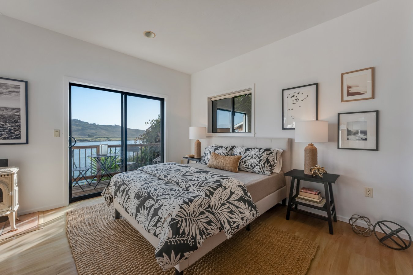 Bedroom with sliding glass door leading to balcony overlooking water, with framed artwork on white walls, a bed with floral bedding, side tables with lamps, and a wooden floor with woven rug.