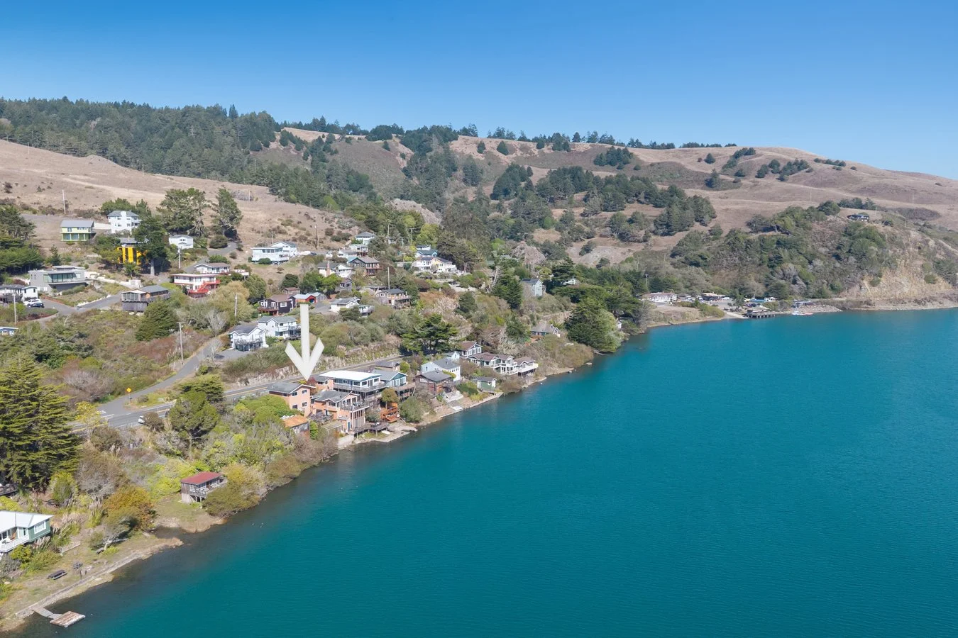 Aerial view of a lakeside residential area with houses along the shoreline and hillside, clear water in the lake, and a hill with trees in the background under a blue sky.