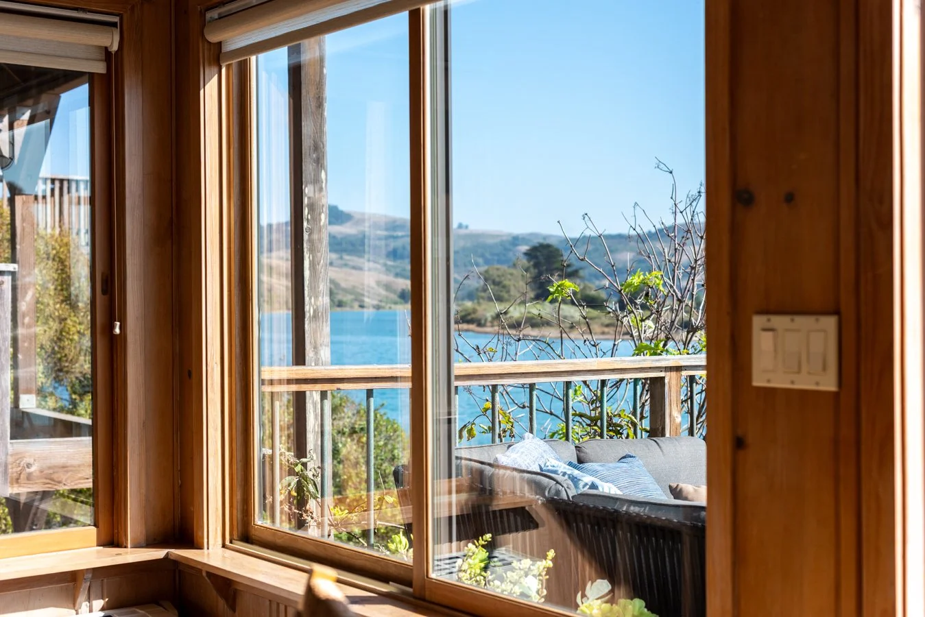 View through wooden-framed sliding glass doors showing a balcony with a sofa and cushions, overlooking a scenic lake and rolling hills under a clear blue sky.