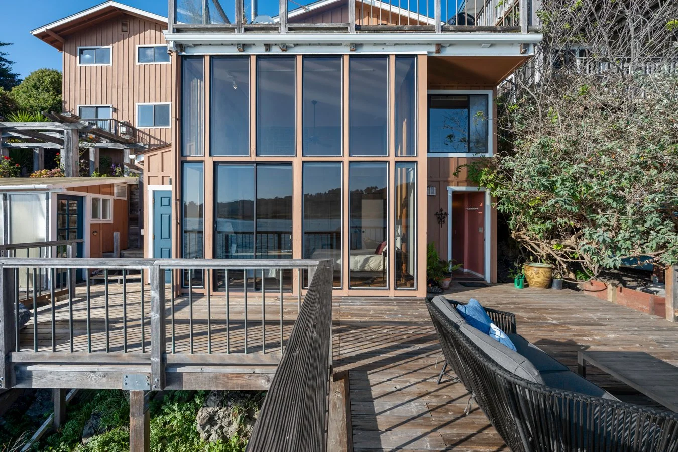 Exterior view of a multi-level house with large glass windows, a wooden deck, outdoor seating, potted plants, and a tree on the right side.