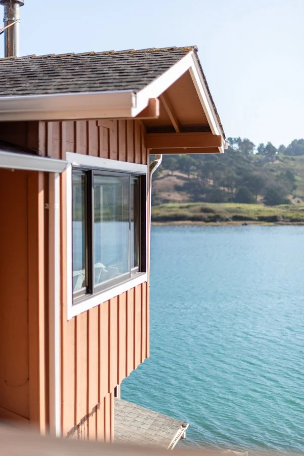 A house with red wooden siding by a body of water with trees and hills in the background.