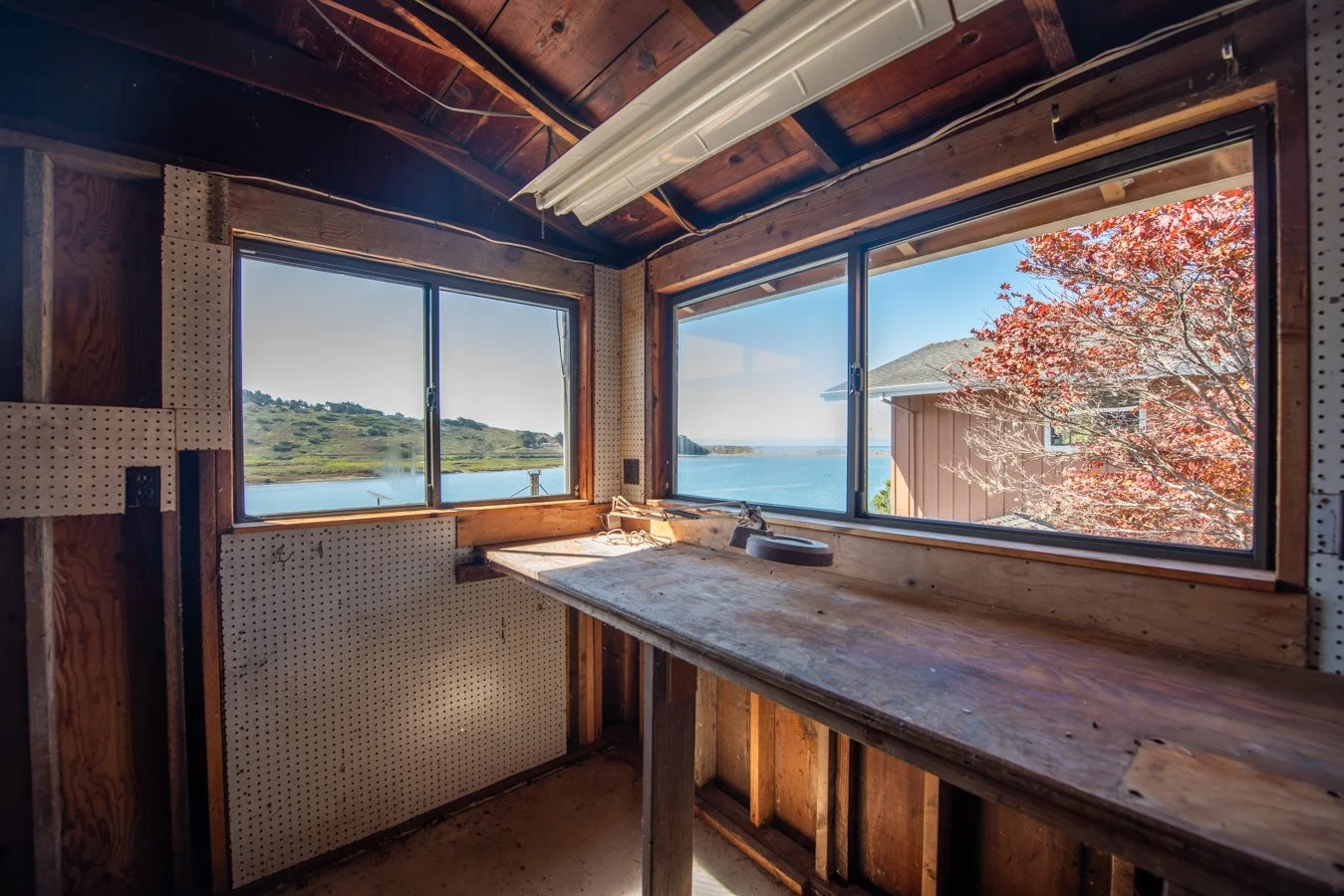 Interior view of a cabin under renovation with a large window showing water, hills, and a tree with red leaves outside.