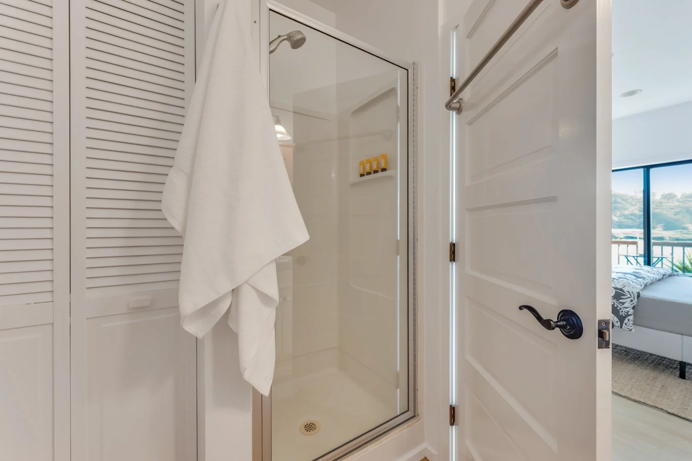 Bathroom shower with a white towel hanging on a bar, a door with a handle, and a view into a bedroom with a bed and large window.