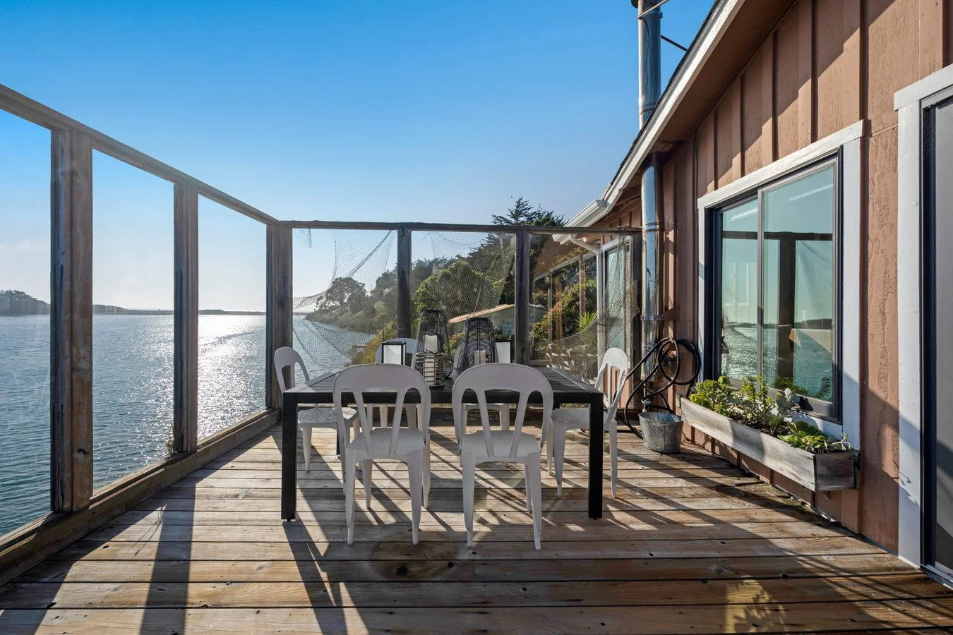 A wooden deck beside a house with a large sliding glass door, overlooking a body of water. There is a black table with white plastic chairs around it, lanterns, and potted plants with sunlight reflecting off the water.