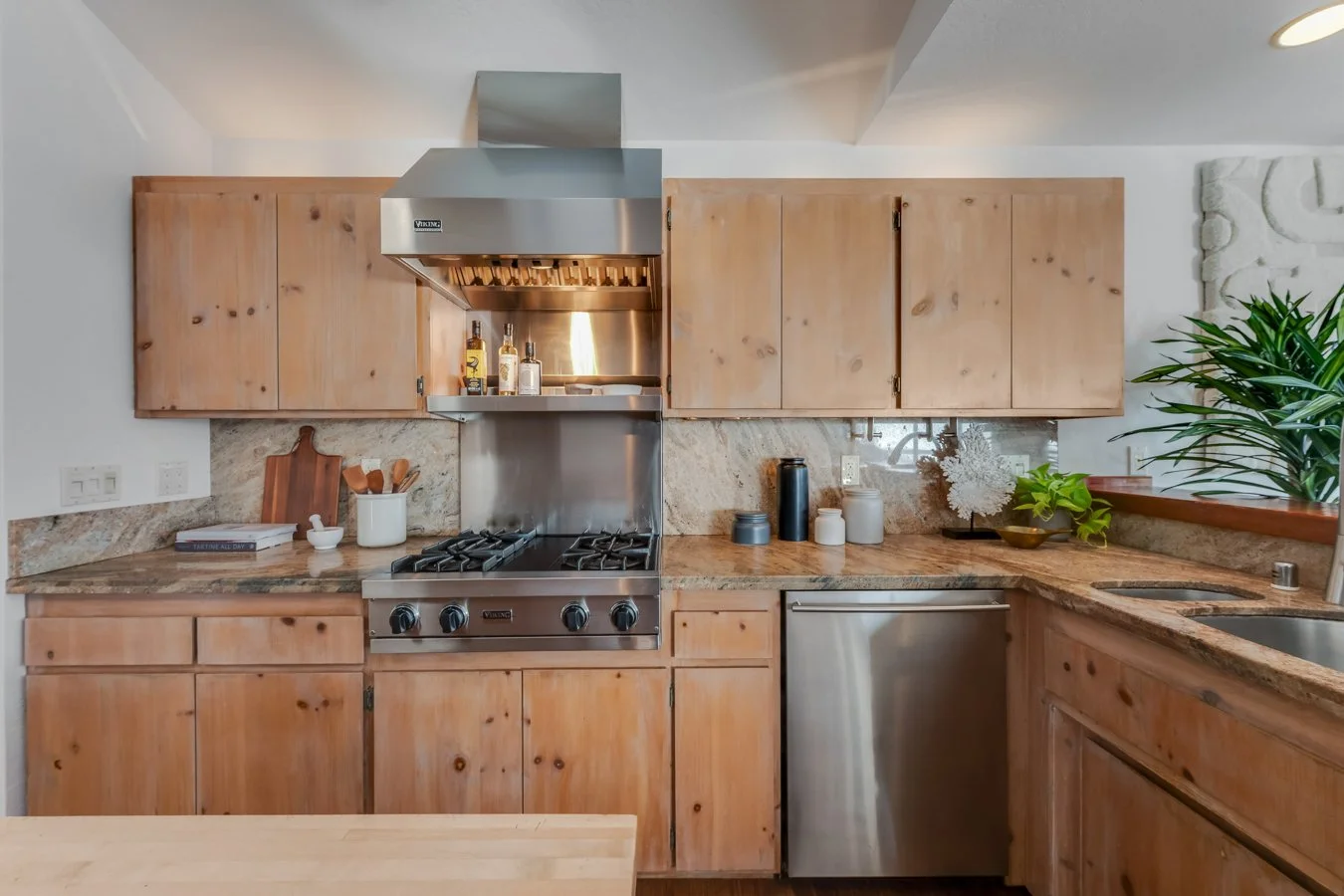 Kitchen with wooden cabinets, stainless steel stove and range hood, granite countertops, and various small plants and accessories. There is a cutting board, bottles, and a book on the counter.