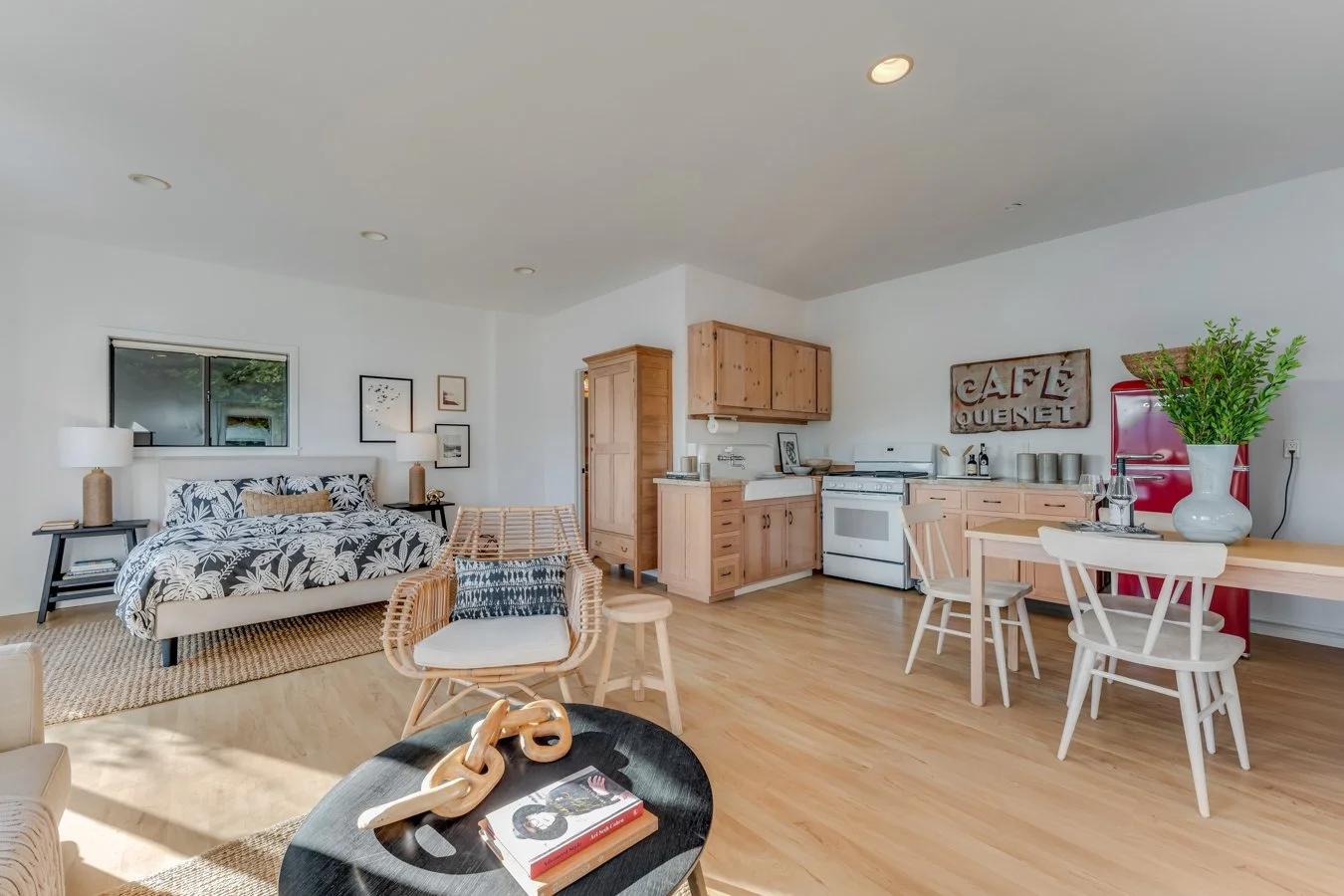 Open-concept living room and kitchen with light wood furniture, a white sofa with black and white floral pillows, wood dining table with chairs, and a sign that says 'Café Queret'.