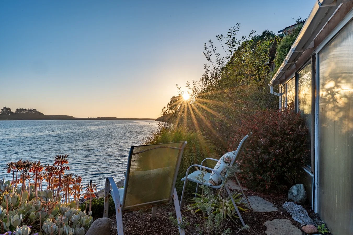 A lakeside outdoor scene at sunset with a house, chairs, and plants along the shoreline.