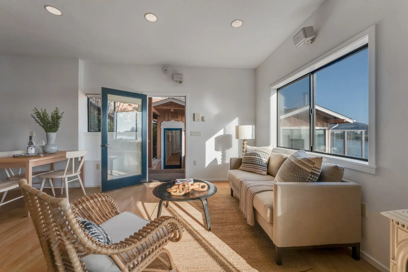 Bright living room with large window, beige sofa with patterned pillows, wicker chair, small round black coffee table with sculptures, wooden dining table with chairs, and door leading outside. Natural sunlight streams in, illuminating the space.