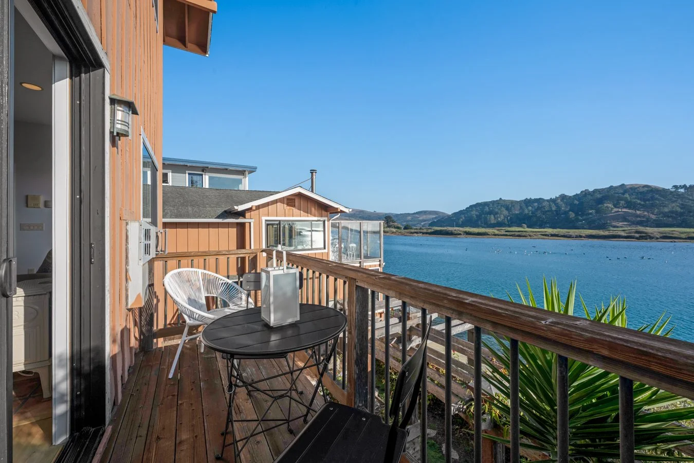 Balcony with black round table, white and black chairs, overlooking water and distant hills under a clear blue sky.