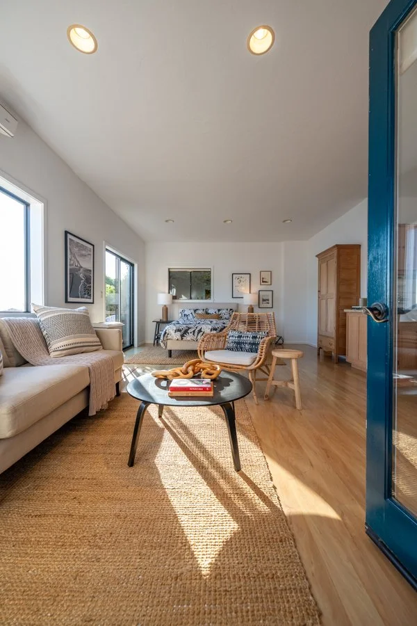 Bright living room with a black coffee table, beige sofa with striped pillows, rattan chairs, a bed in the background, and large windows letting in natural light.
