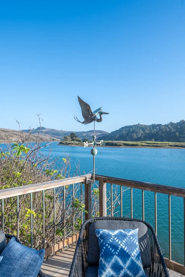Patio with a chair and pillow overlooking a lake with hills in the background, clear blue sky, and a weather vane featuring a heron.