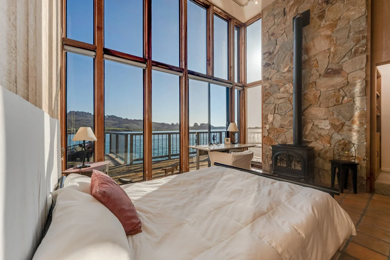 A cozy bedroom with large floor-to-ceiling windows overlooking a body of water and hills, featuring a bed with white linens and a pink pillow, a bedside table with a lamp, a stone fireplace, a desk with a chair, and a balcony outside.