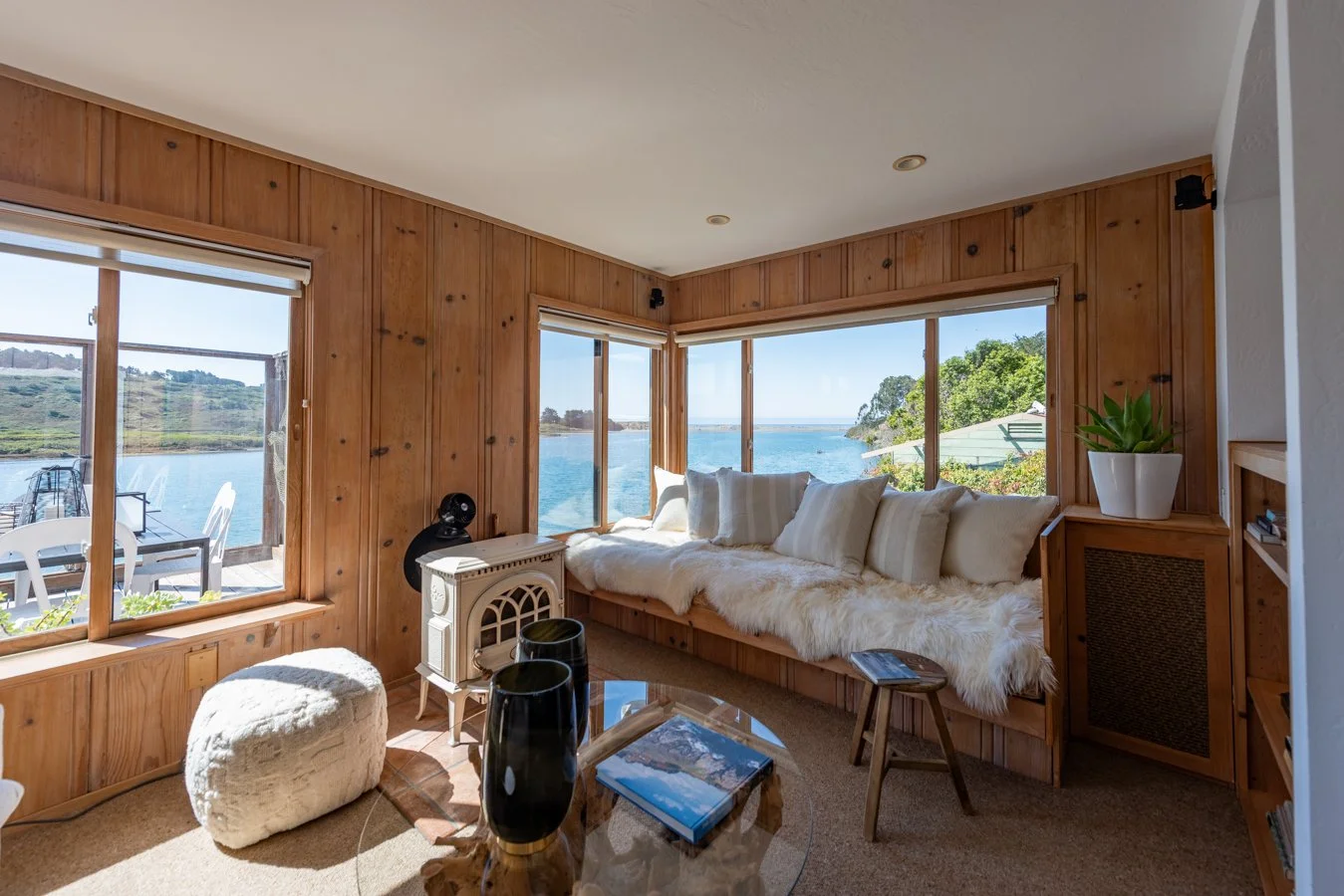 Living room with large windows overlooking a body of water, featuring a built-in wooden window seat with white cushions and fur throw, a glass coffee table, and a potted plant on a shelf.
