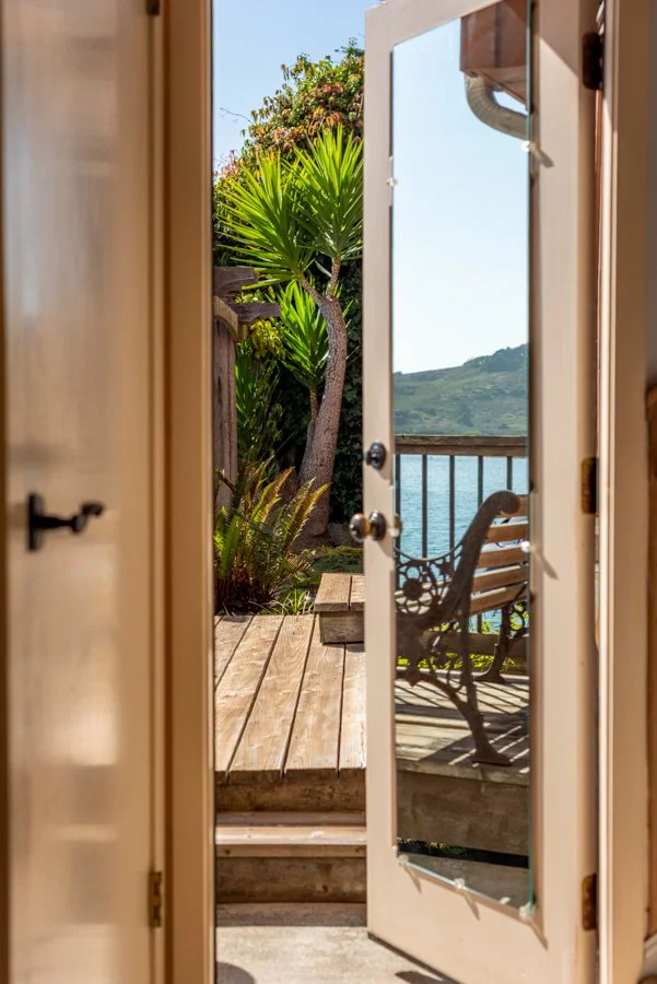 View through a glass door onto a wooden deck with outdoor bench, overlooking a body of water and green hills in the distance, with lush plants and a tree nearby.