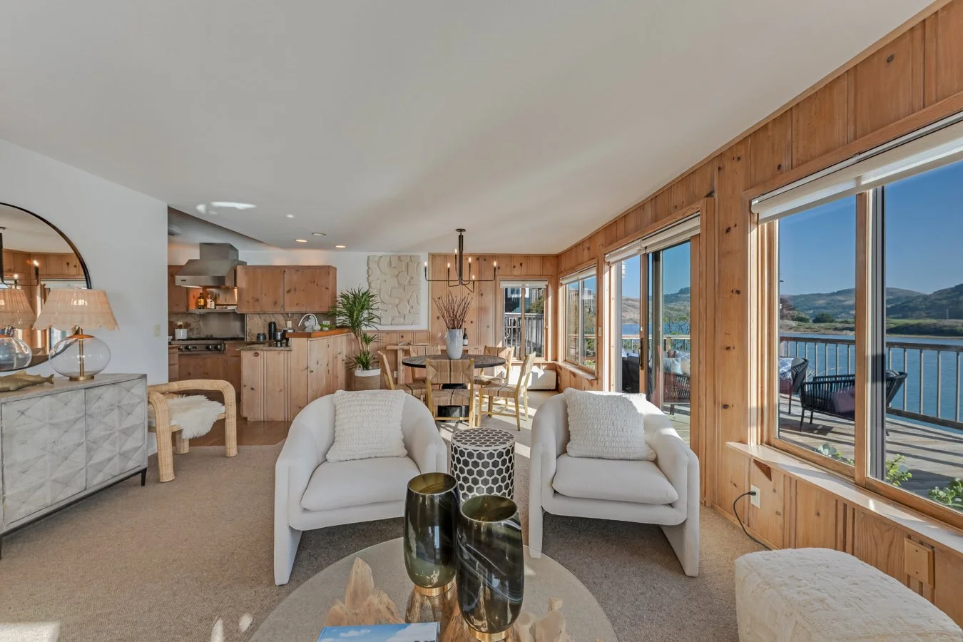 Living room with large windows overlooking a lake and mountains, featuring white armchairs, a round coffee table with vases, a dining table with chairs, and a kitchen area with wooden cabinets.