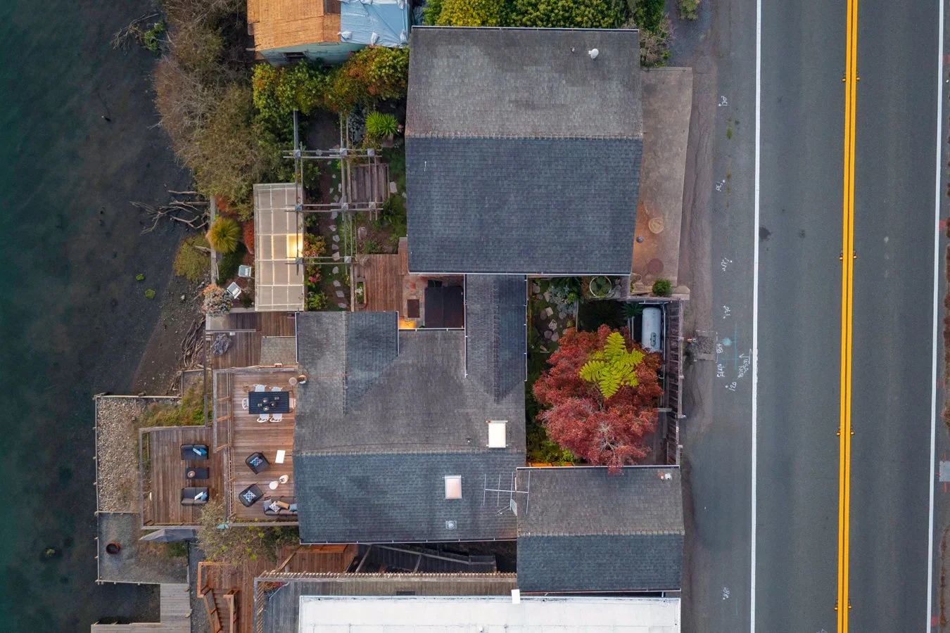 Aerial view of a residential backyard with a deck, outdoor furniture, trees with red and green leaves, and neighboring houses, next to a street with double yellow lines.